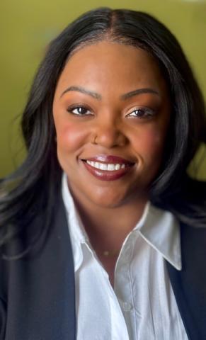 A dark-skinned black woman with long dark hair and a white shirt under a black blazer.