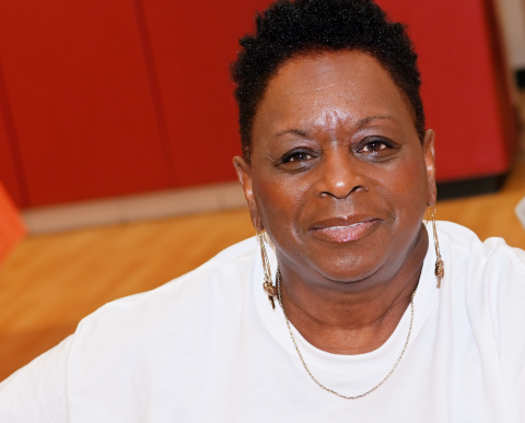 A dark-skinned woman with short dark hair smiles in front of a red background. She is wearing a white shirt and gold jewelry.