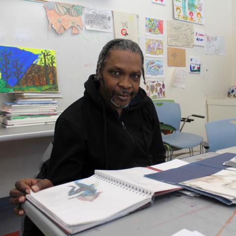 A dark-skinned man with short braids sits at a table with his artwork spread out.
