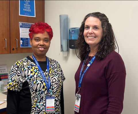 A dark-skinned woman with red hair wearing scrubs and a light-skinned woman with brown hair wearing a sweater.