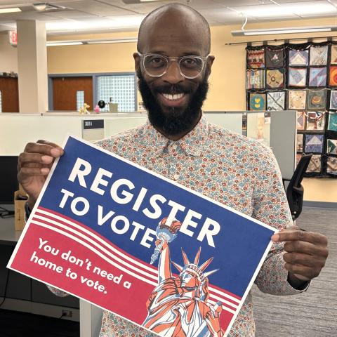 Dark-skinned man in an office holding a blue and red sign that reads “Register to vote” and “You don’t need a home to vote.” The sign includes an illustration of the Statue of Liberty and red stripes.