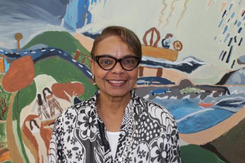 A medium-skinned woman wearing glasses and a floral black and white blouse smiles in front of a colorful painting