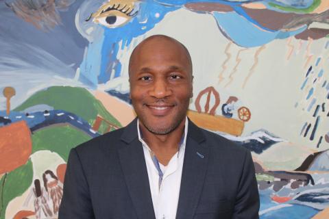 A dark-skinned man wearing a suit smiles in front of a colorful painting.