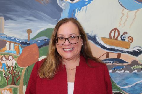 A light-skinned woman wearing glasses and a red blazer smiles in front of a colorful painting
