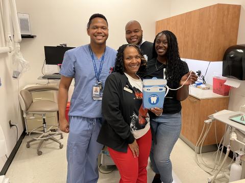 Four adults pose in a dental suite, smiling. The woman in front holds an oversized dental floss.