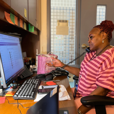 A dark skinned person speaking on the phone while sitting at their computer.
