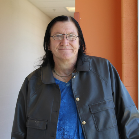 A light-skinned person with dark shoulder length hair and glasses, wearing a blue shirt and black jacket.