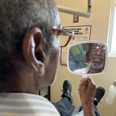 A dark-skinned man with grey hair and glasses looks at his teeth in a hand mirror.