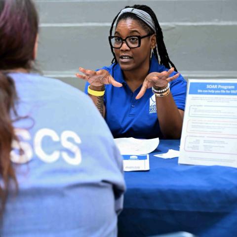 A dark-skinned woman with long dark hair behind a table speaks to an incarcerated woman.