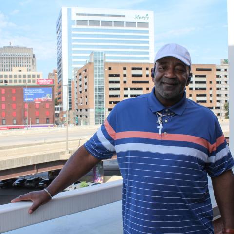 A dark-skinned man with a white baseball hat and blue shirt with the Baltimore skyline behind him.
