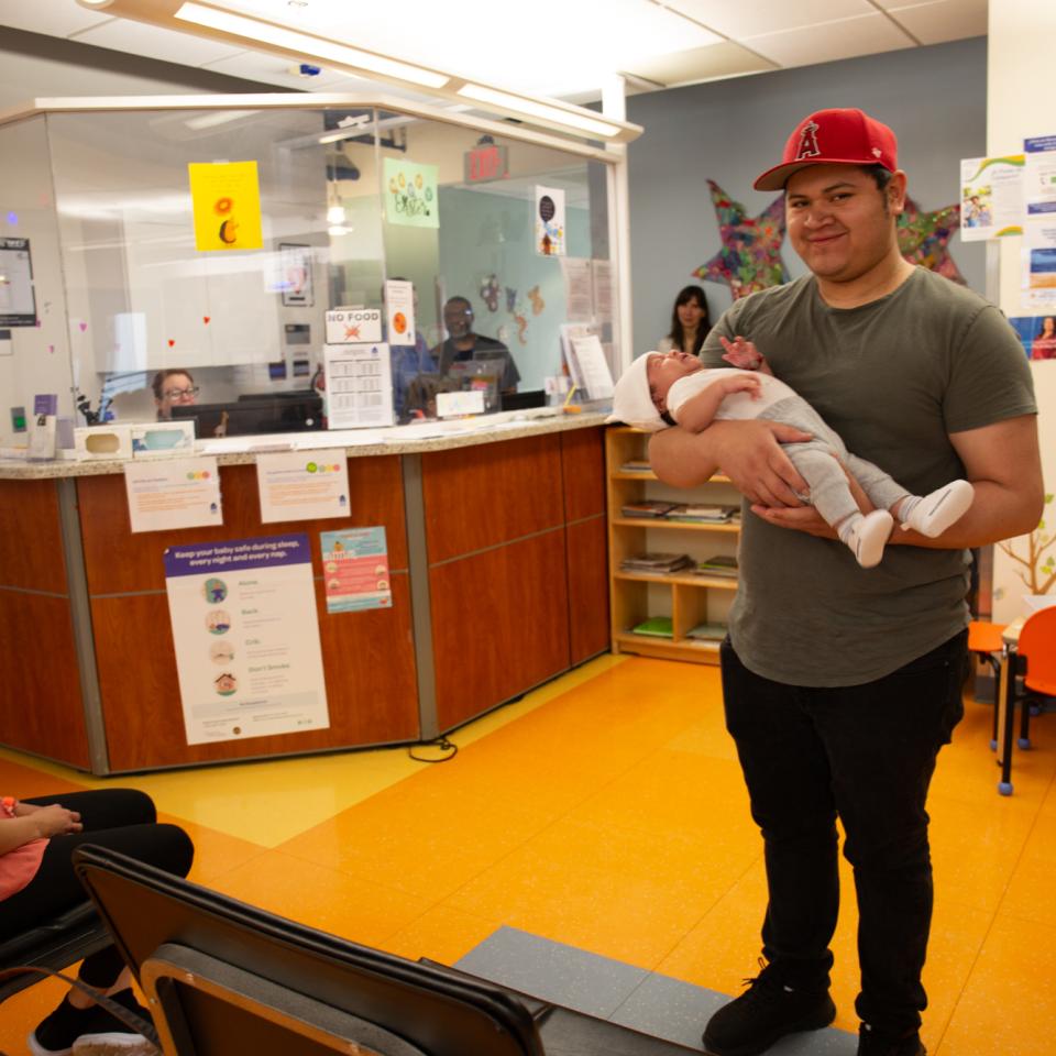 A man wearing a red baseball cap stands in a clinic waiting area cradling his small child