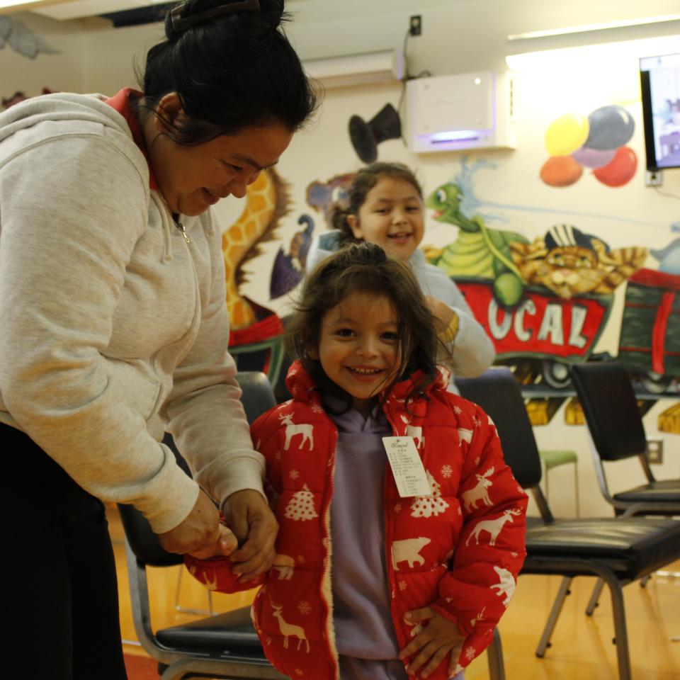 A young girl with brown hair wears a red coat and holds her mom's hand.
