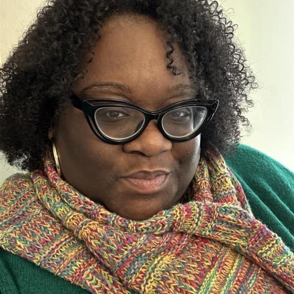 A dark-skinned woman with short curly hair wearing black-framed glasses and a colorful scarf.