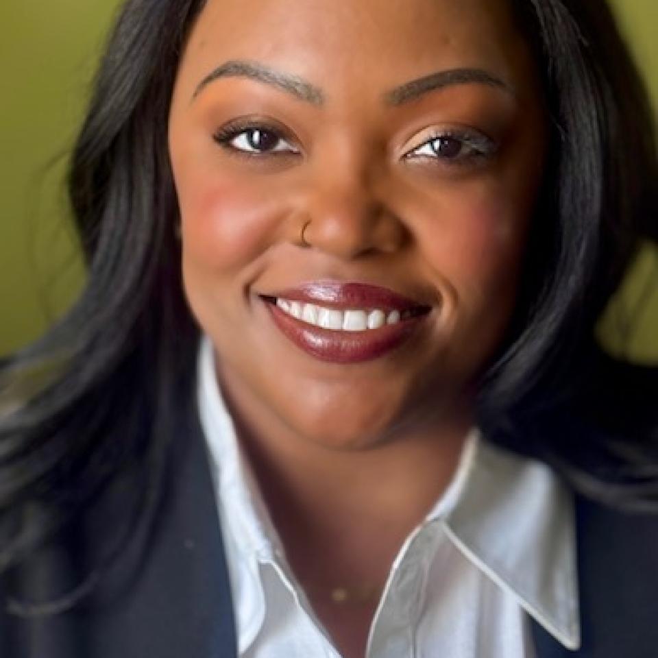 A dark-skinned black woman with long dark hair and a white shirt under a black blazer.
