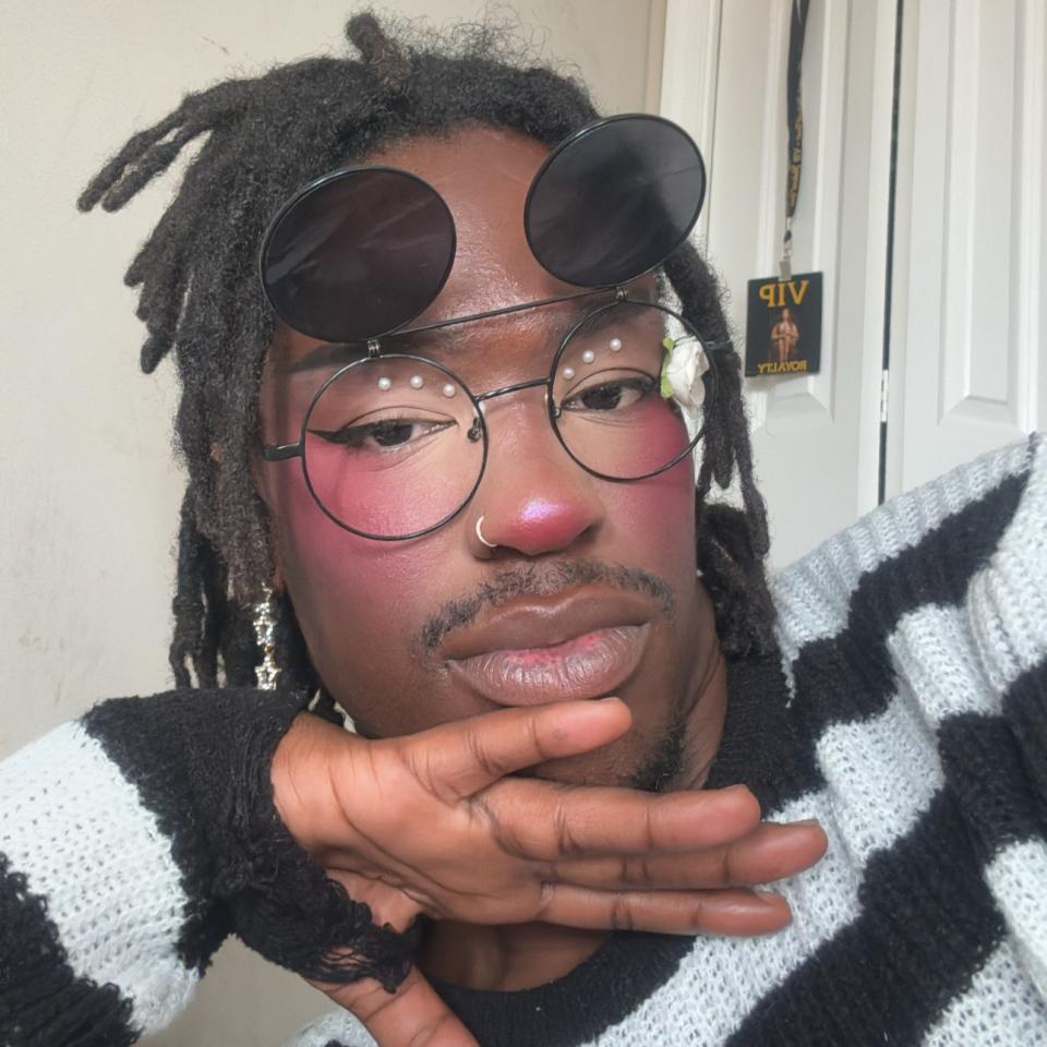 A dark-skinned person with short dark locs wears glasses with flipped up clip-on sunglasses and a stripped sweater.