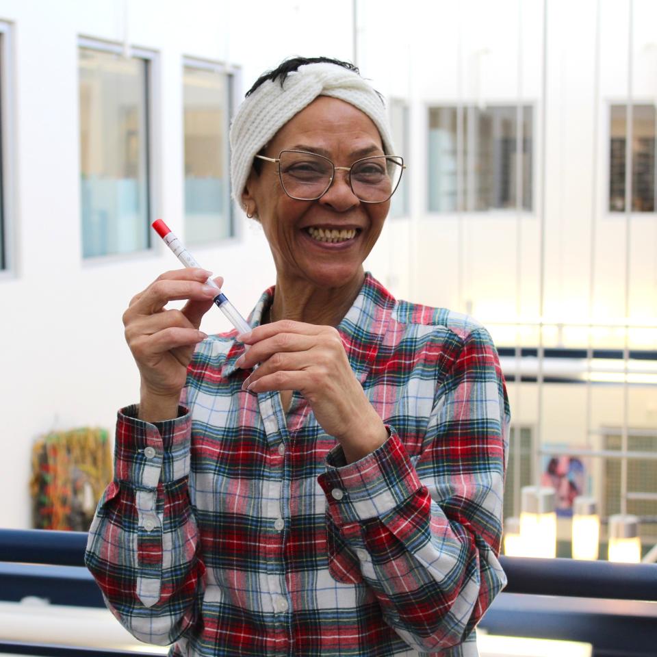 A medium-skinned woman smiles while holding an HPV self-collection test tube. She wears glasses and a head wrap.