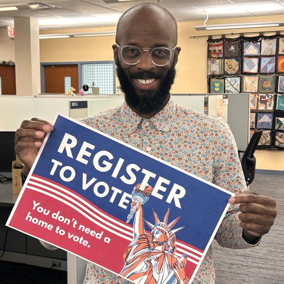 Dark-skinned man in an office holding a blue and red sign that reads “Register to vote” and “You don’t need a home to vote.” The sign includes an illustration of the Statue of Liberty and red stripes.