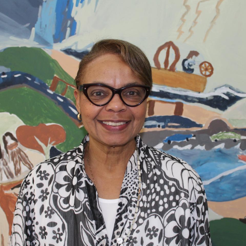 A medium-skinned woman wearing glasses and a floral black and white blouse smiles in front of a colorful painting