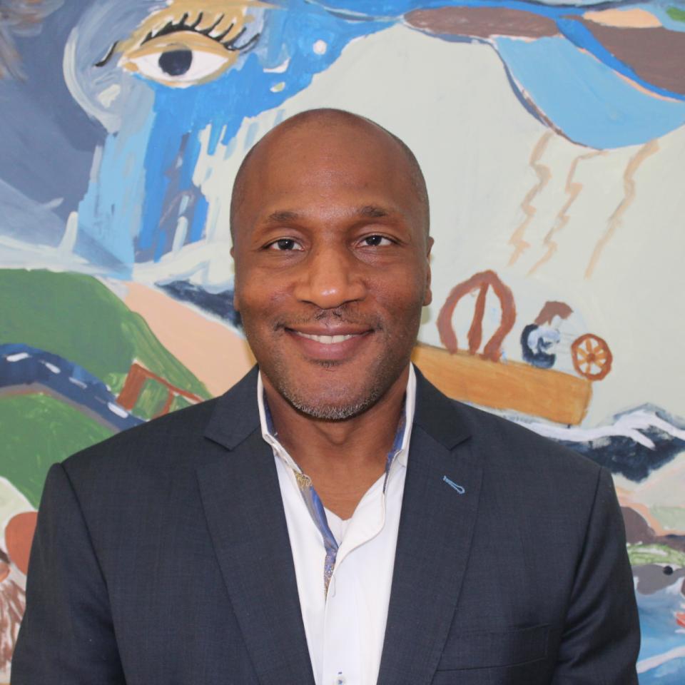 A dark-skinned man wearing a suit smiles in front of a colorful painting.