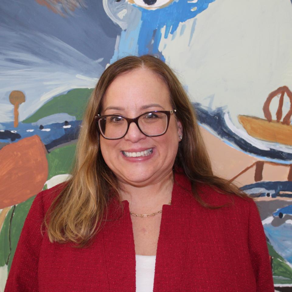A light-skinned woman wearing glasses and a red blazer smiles in front of a colorful painting