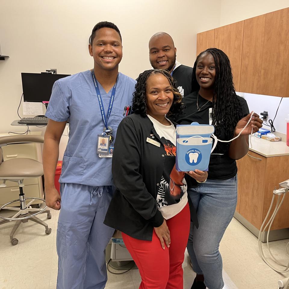 Four adults pose in a dental suite, smiling. The woman in front holds an oversized dental floss.