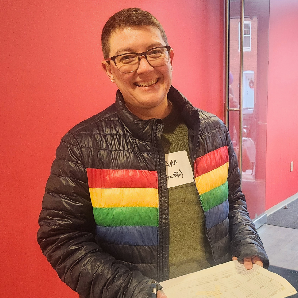 A light-skinned person with short brown hair and glasses wearing a brown puffy coat with rainbow stripes.