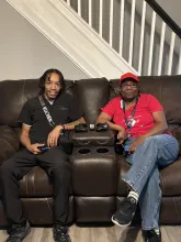 A dark-skinned man man with short dark braids sits on a brown couch next to a dark-skinned woman wearing a red hat.