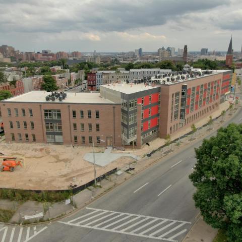 A high view of the entire Sojourner at Oliver complex, with brick front exterior walls, construction vehicles in a sandy lot, and trees/a view of Baltimore in the background. 