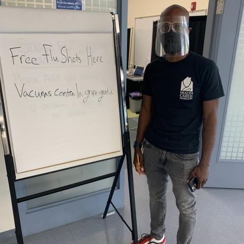 Malcolm Williams, Client Relations Coordinator, wearing a Health Care for the Homeless shirt, face mask, and face shield, in front of a sign reading 