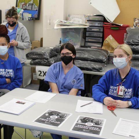 Several nursing students from Notre Dame of Maryland with varying skin tones look at the camera, all wearing blue scrubs, sweatshirts, and facemasks, sitting at a table. Plastic wrapped coats are seen behind them. 