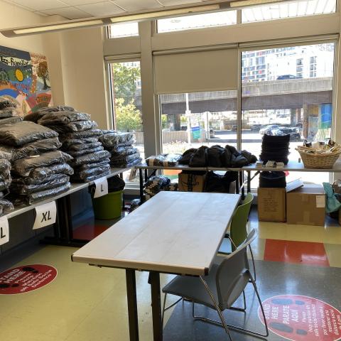 An empty registration table and tables full of unopened, plastic-wrapped coats sit in an empty, clinical room with bright sunlight coming in through the windows.  The coats are organized and labeled with 